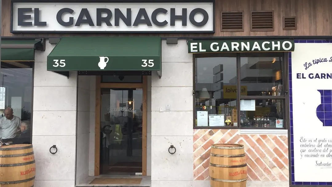 Fachada de La Típica Bodega El Garnacho: rótulo, toldos verdes con la jarra y barricas en la puerta